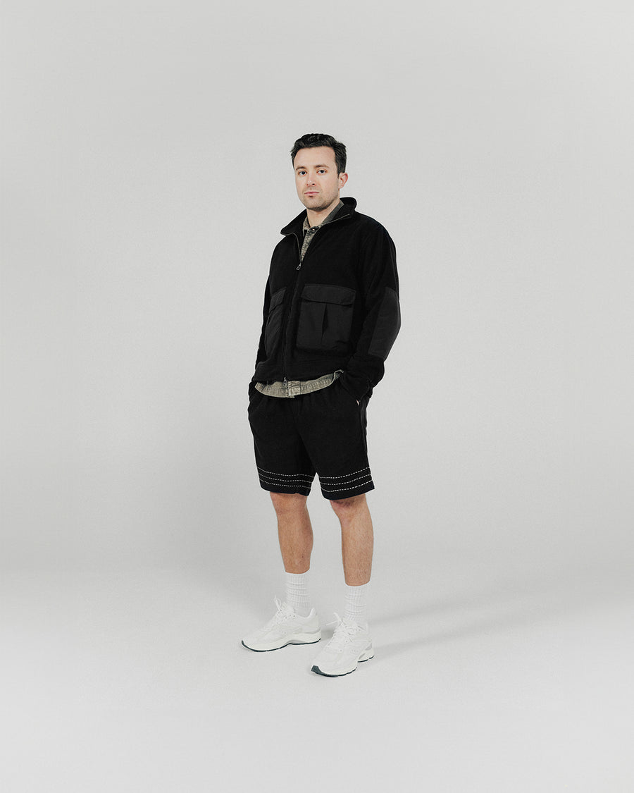 A man poses in a black fleece vest and shorts with white sneakers.